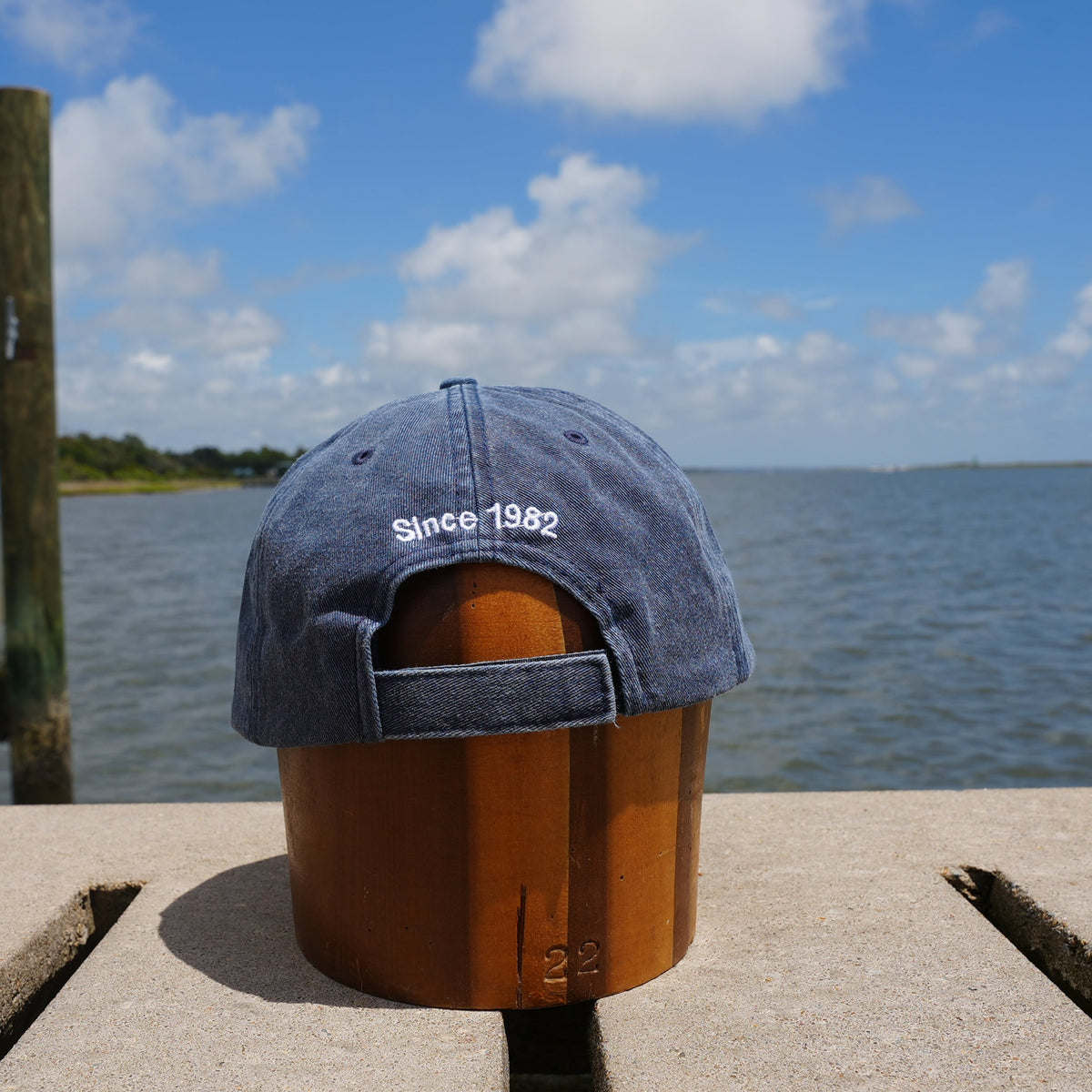 A blue denim baseball cap with 'Since 1982' embroidered on the back, displayed on a wooden stand on a concrete dock with a lake and sky in the background.