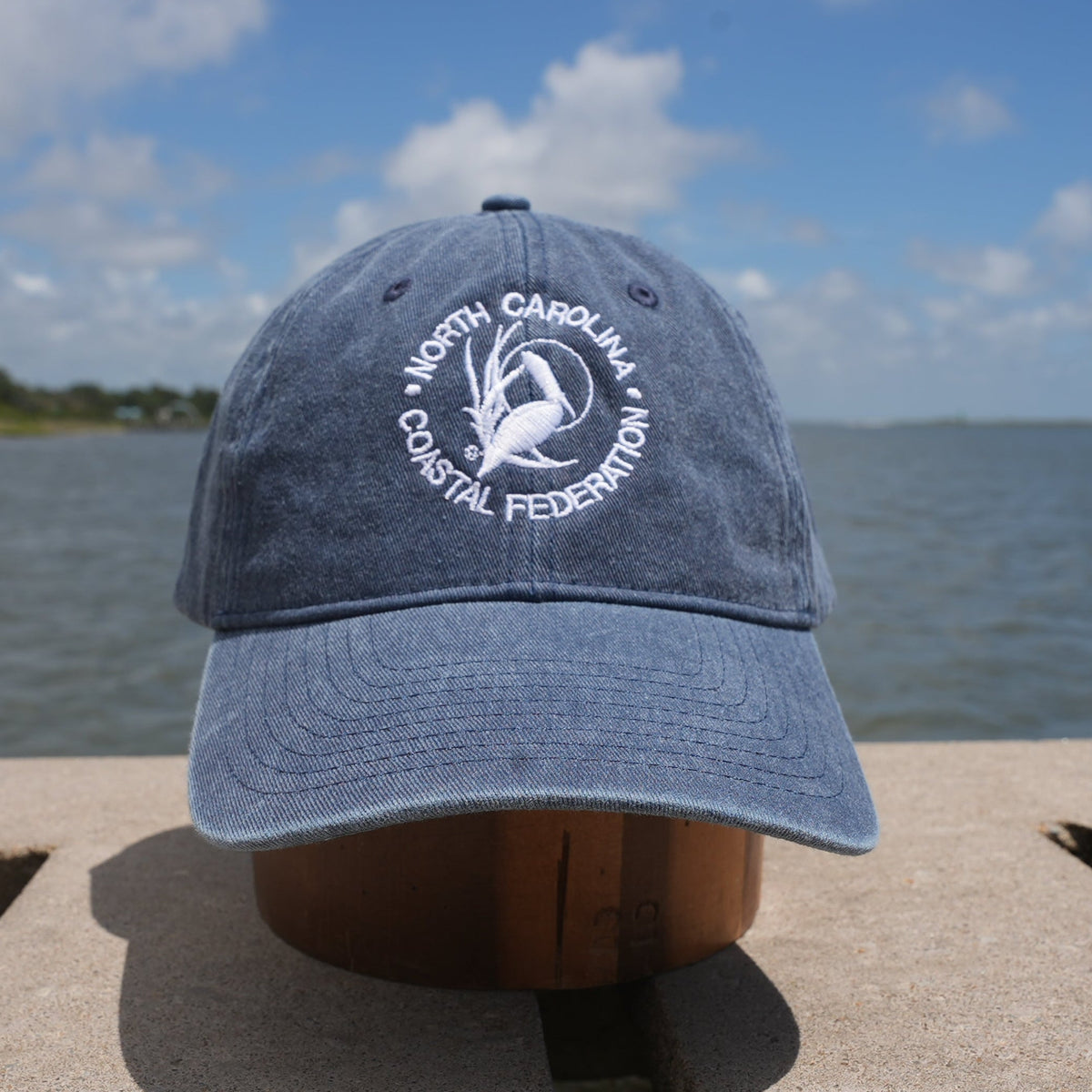 A blue baseball cap with a white embroidered logo for the 'North Carolina Coastal Federation' sits on a wooden dock with water and a cloudy sky in the background.