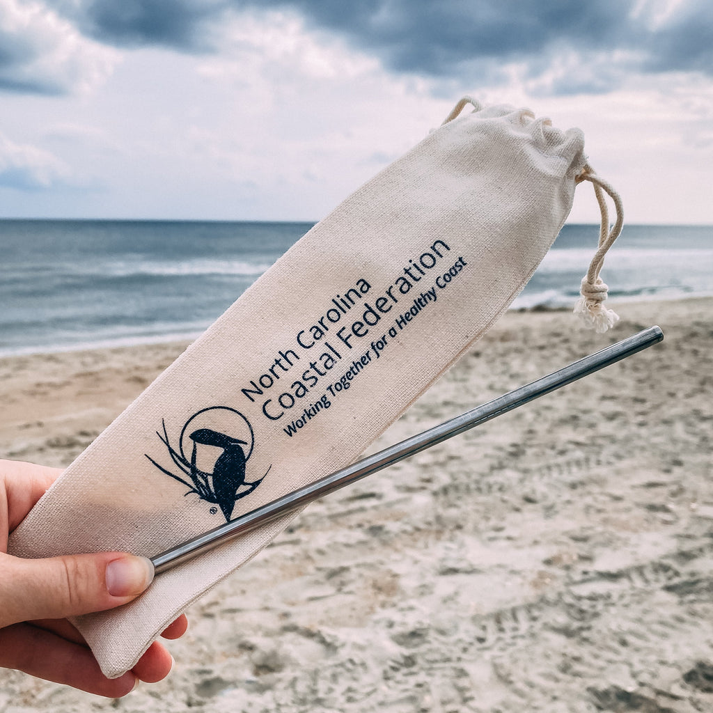 Straw and drawstring bag with North Carolina Coastal Federation logo on a beach.