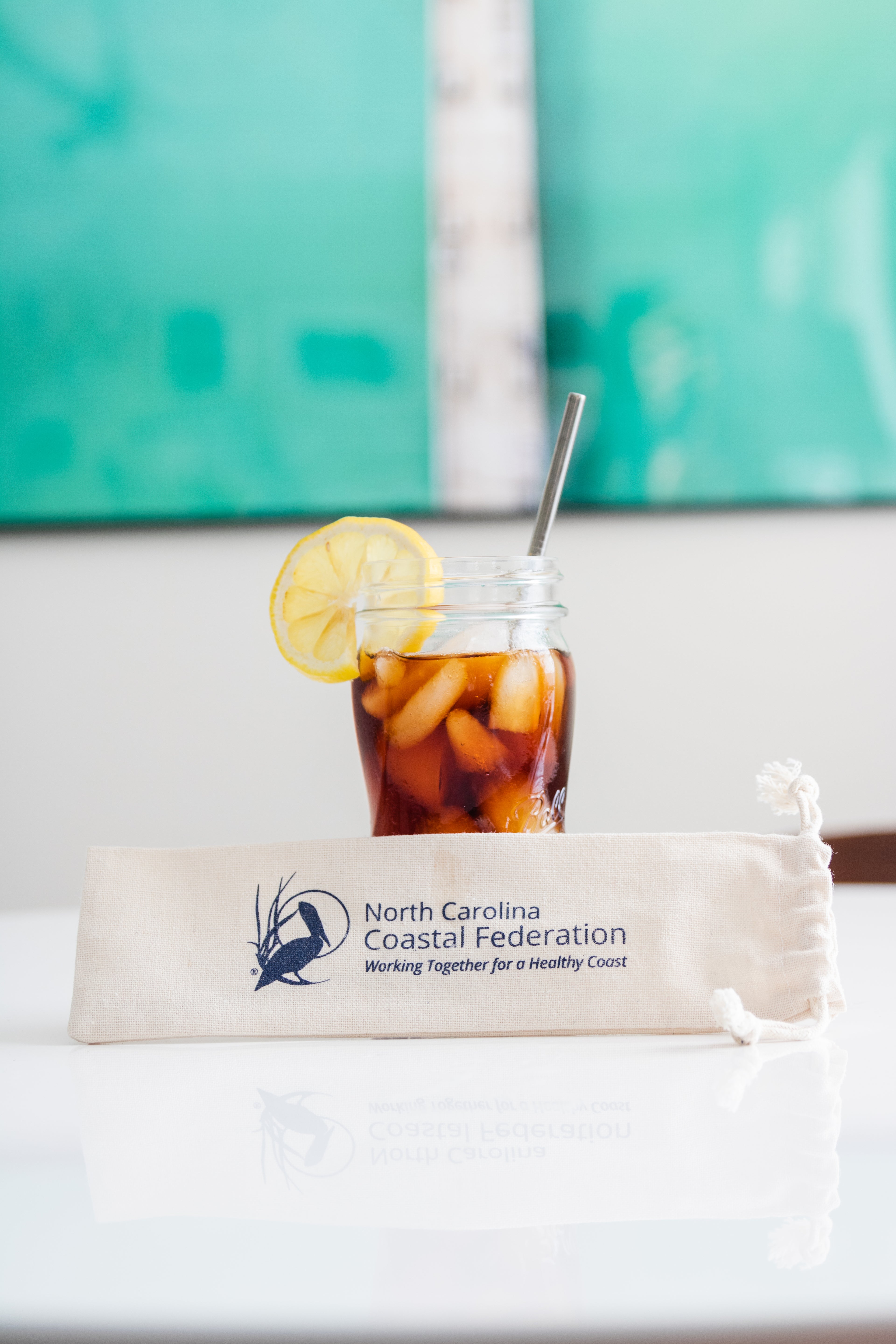 Iced drink with a lemon wedge and straw in a mason jar, placed on a white surface with a North Carolina Coastal Federation bag underneath.